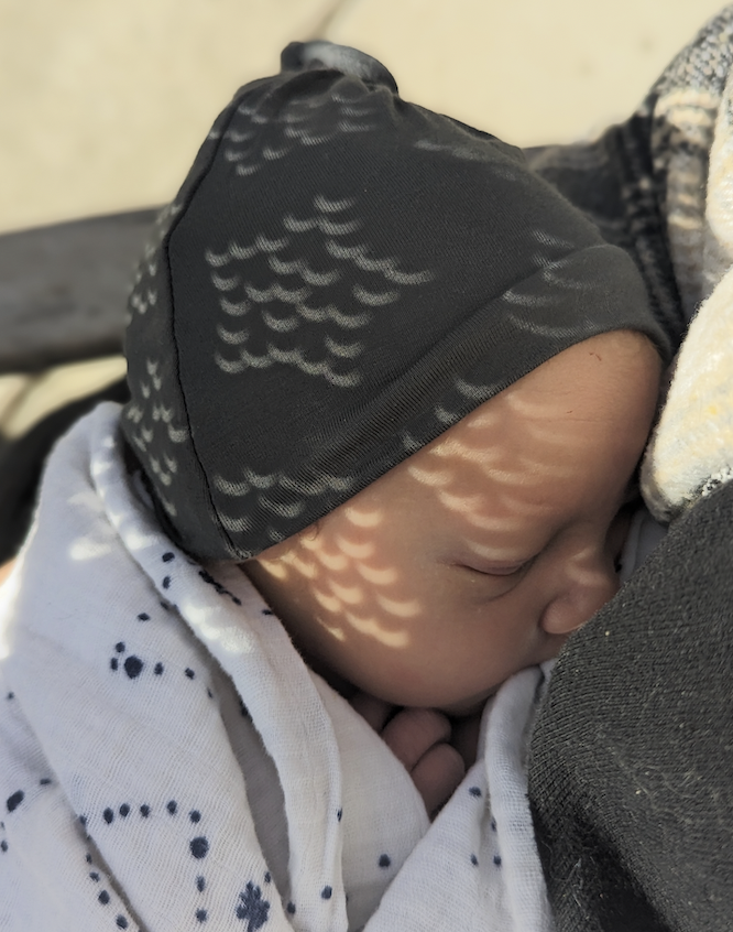 Image of my son as a one-day-old with small crescent lights on his face from the eclipse in 2024. He is wearing a grey hat and has a black and white stars swaddle on.